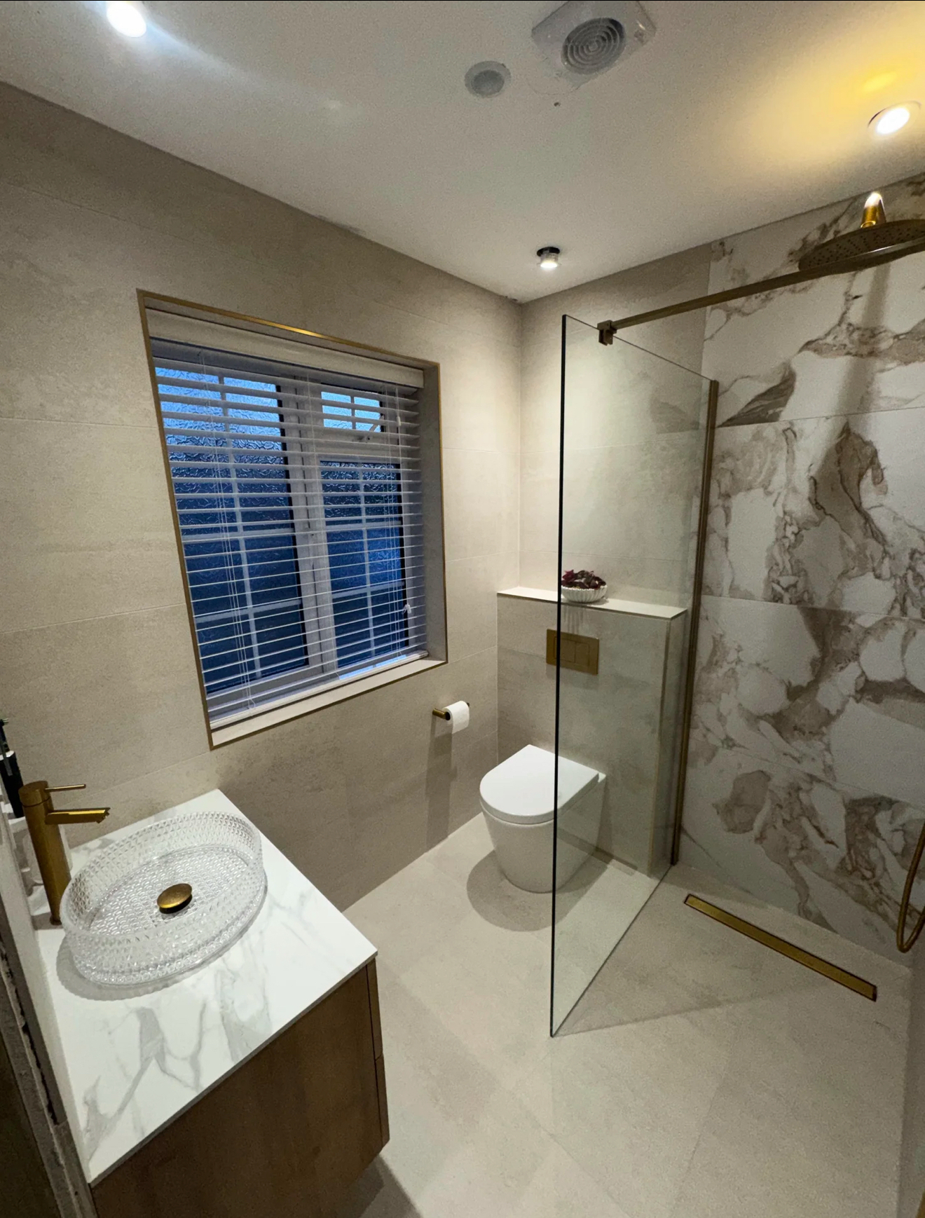 Elegant marble bathroom with clear glass walk-in shower, vessel basin, brass tap and window shutters